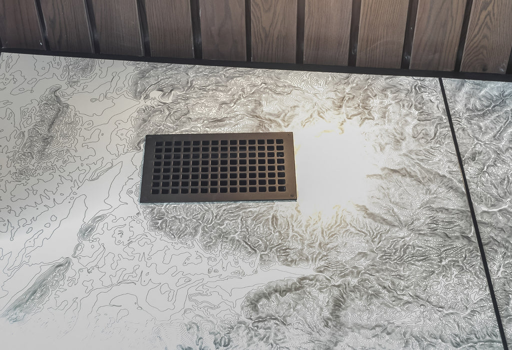 Cast aluminum vent cover installed in a wall with topo map wallpaper with wood slat ceiling.