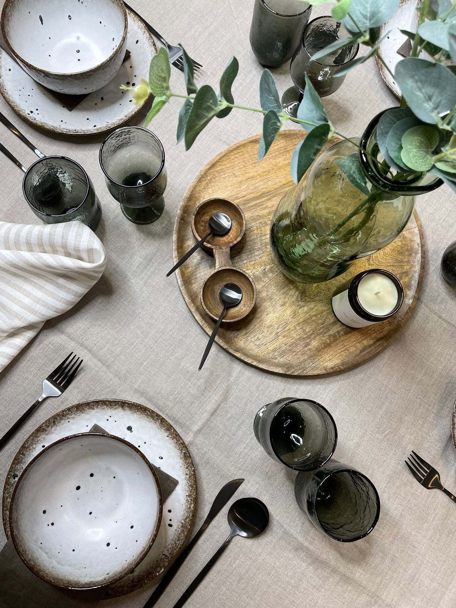table setting with beige tablecloth, grey cutlery and glasses, white and brown plates and bowls and a central round wooden board with a green glass vase and foliage, a candle and empty small wooden bowls