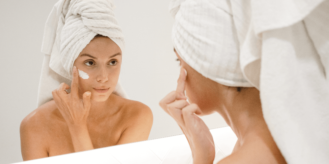 Image depicts a woman wrapped in a towel, with another towel turbaned around her head, looking into a mirror as she applies tretinoin cream to her face. She has a slight smile, indicating a moment of self-care and satisfaction. The neutral, white background enhances the clean, calm atmosphere typical of a skincare routine setting.