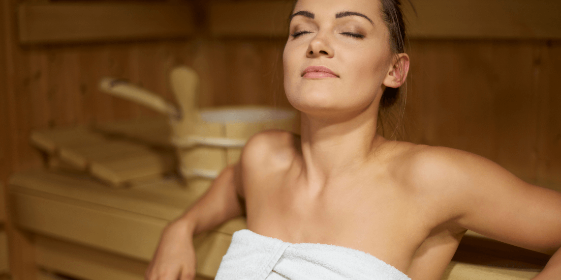 Image shows a woman in a sauna with her eyes closed, exuding a sense of relaxation and contentment. She is seated comfortably, surrounded by the warm wooden interior of the sauna. The calm expression on her face and the serene sauna setting convey a peaceful, restful moment.