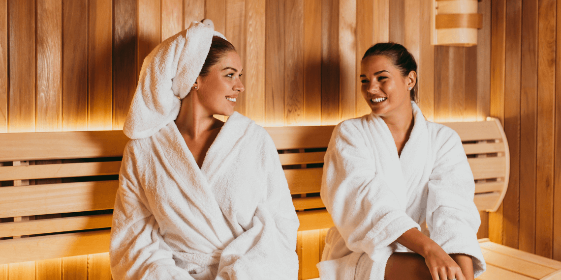 Image shows two friends enjoying a relaxing moment in a sauna. They are seated side by side on wooden benches, smiling contentedly. The warm, amber tones of the sauna enhance the serene and comfortable atmosphere, highlighting the camaraderie and relaxation shared between the two individuals.