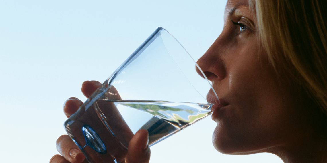 Image captures a woman sipping water from a glass, set against a clear blue sky. She looks refreshed and hydrated, emphasizing the importance of drinking water. The bright daylight and blue backdrop highlight her relaxed demeanor and the healthy action of hydrating.
