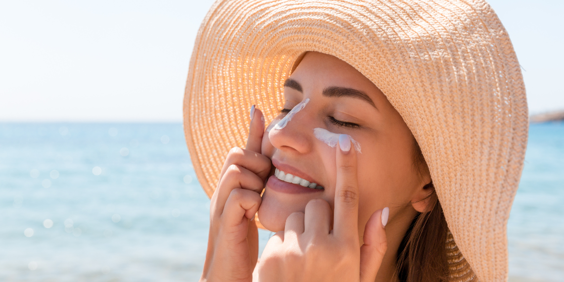 Image shows a woman at the beach applying sunscreen to her cheeks, smiling as she faces the sun. Her joyful expression and the act of applying sunscreen emphasize the importance of skin protection while enjoying the outdoors. The sunny beach setting enhances the vibrant, healthy outdoor activity.