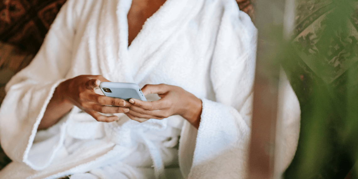 Image features a woman in a white robe, intently searching through FAQ questions on her smartphone. The background is softly blurred, conveying an aesthetic reminiscent of a tranquil spa setting. This serene atmosphere enhances the focus on the woman, who appears engaged and thoughtful as she interacts with her device.