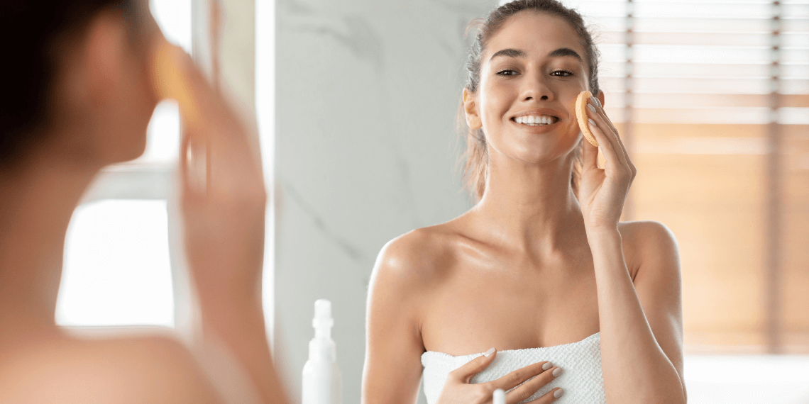 Image captures a woman smiling at her reflection in a mirror as she applies skincare products to her face using a pad or cloth. Her skin glistens, enhanced by the skincare routine, reflecting the light and highlighting her radiant complexion. This moment of self-care is framed in a personal and intimate setting, emphasizing her enjoyment and dedication to maintaining her skin's health.