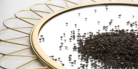 Close-up image of a decorative gold tray with a mirrored surface, on which Bakuchiol seeds are scattered. The seeds reflect in the mirror, emphasizing their natural texture and the elegant setting of the tray. The composition highlights the luxurious and decorative presentation of the natural skincare ingredient.