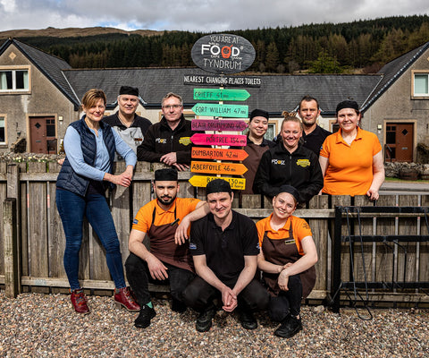The Real Food cafe team next to a signpost showing how far are nearest changing places toilets from Tyndrum, Spend a Penny Sotland campaign