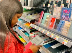 African-American woman in red long-sleeved top with long dark brown hair picking up Achiever planner in store with several other planners on shelves