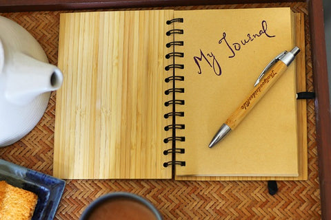 Open wood-grain covered journal with "My Journal" written on first page of brown paper. There is a woodgrain and silver pen sitting on first page. Journal is istting on multi-shade tan placemat. There is a white ceramic teapot, blue cup of tea on the placemat.