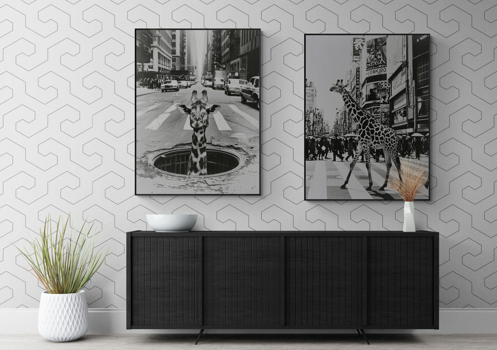 L'image présente un intérieur élégant avec un buffet moderne noir, surmonté de deux photographies en noir et blanc accrochées au mur, l'une montrant une girafe émergeant d'une bouche d'égout, l'autre une girafe traversant un passage piéton, le tout contrastant avec le motif géométrique du papier peint et agrémenté d'une plante verte dans un pot blanc texturé.