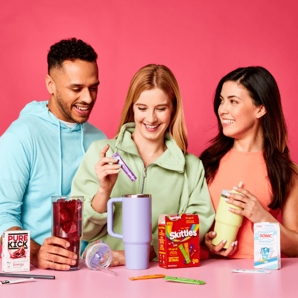 Three friends enjoying a moment together with drink mixes. One person is preparing a drink with a Pure Kick Energy packet, another is holding a Skittles box beside a blender, and the third is sipping from a Sonic branded cup, all smiling and engaged in a fun, casual setting with a pink background