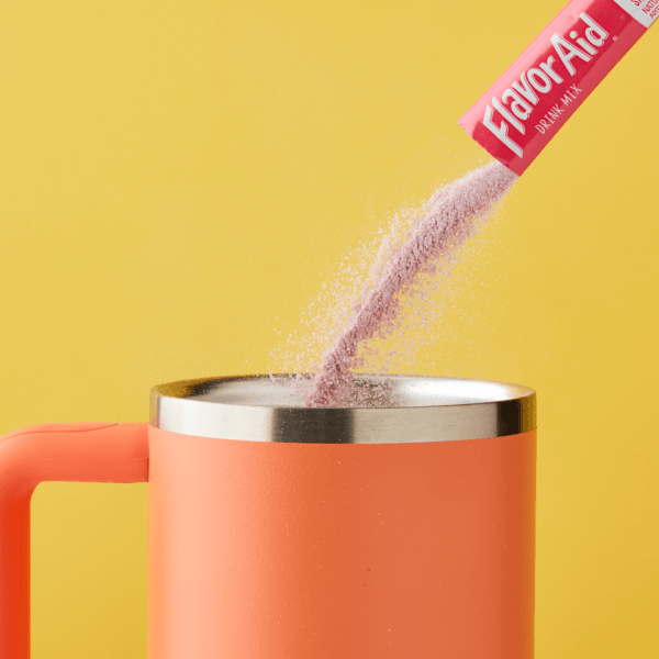 A close-up of a pink Flavor-Aid drink mix being poured into a coral-colored insulated tumbler against a vibrant yellow background, capturing the dynamic motion of the powder as it cascades into the liquid