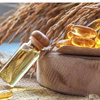 A close-up image showing a small glass bottle with a wooden cap, filled with a golden liquid, likely an oil, and golden capsule supplements scattered around, all set against a background featuring wheat stalks, suggesting a theme of natural ingredients and health supplements.