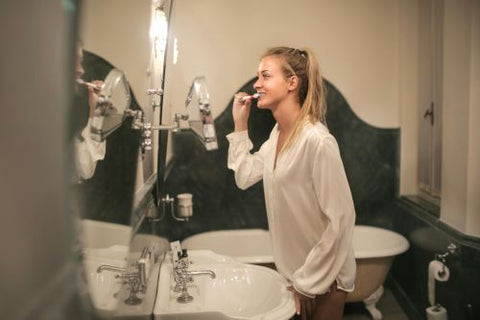 Une femme en chemise blanche se tient devant sa glace dans la salle de bain et se lave les dents
