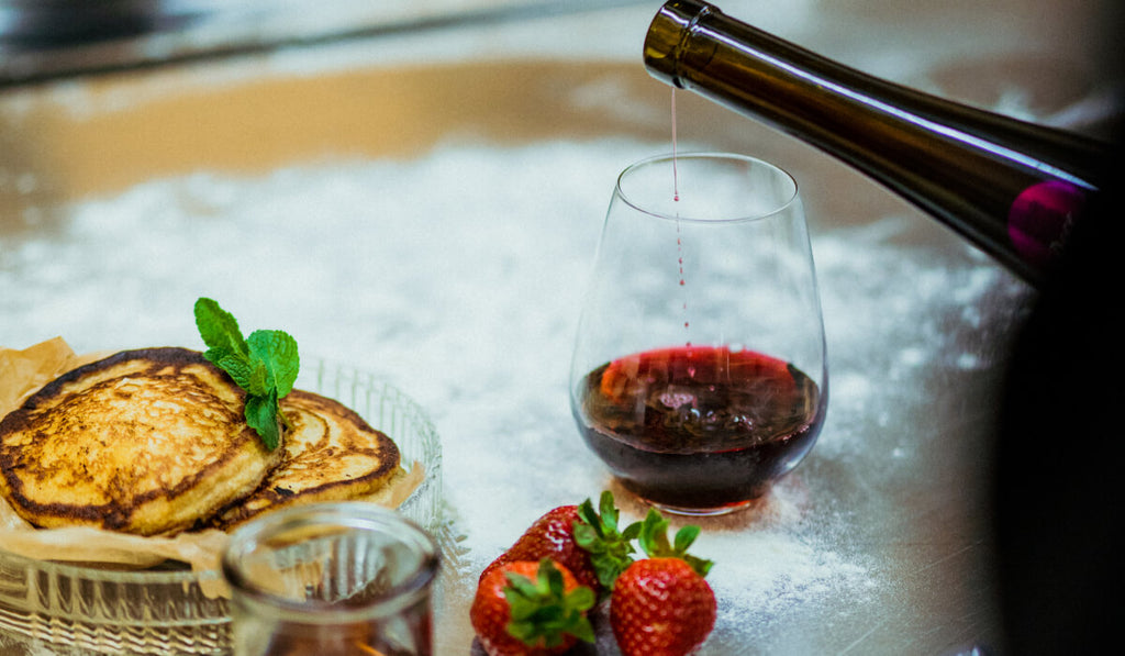 Ein Glas in das Dessertwein geschenkt wird, davor ein Teller mit Pancakes, frische Erdbeeren und Ahornsirup