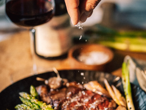 Fingers with a pinch of salt over asparagus, meat next to glass of red wine