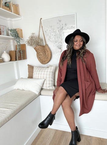 indian woman sitting with legs crossed wearing black hat, brown oversized cardigan with pockets and a black turtleneck dress with black booties.