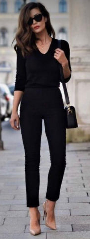 Brunette woman standing on the sidewalk wearing sunglasses, a purse and an all black outfits with nude pumps