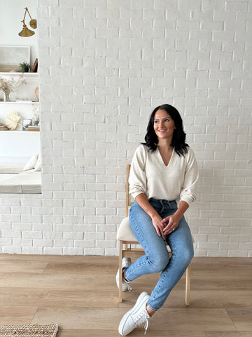 brunette woman sitting on chair wearing cream v neck sweater and jeans with white sneakers