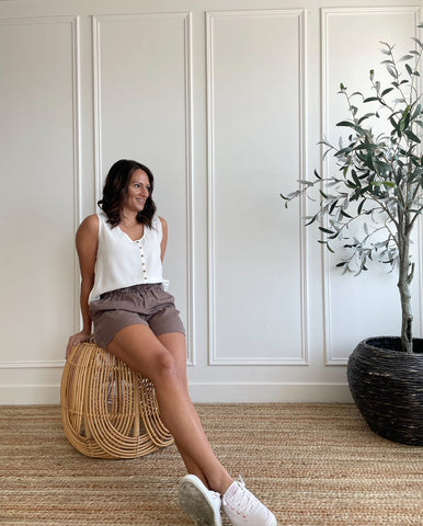 woman wearing white tank top and paperbag shorts with white sneakers sitting on a wicker stool