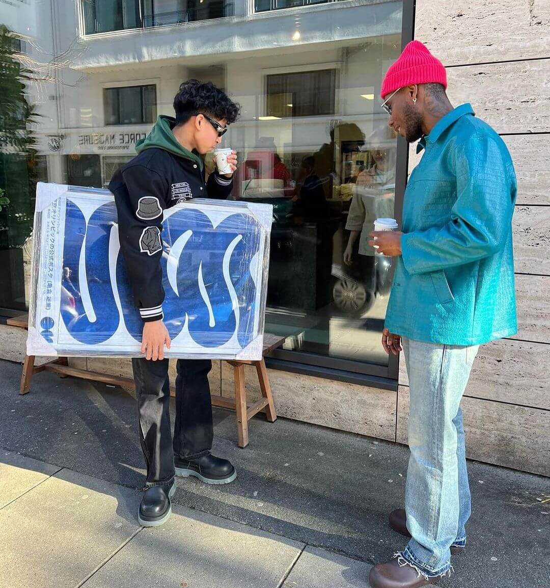 2 men standing on the pavement drinking coffee with one holding a large framed artwork under his arm