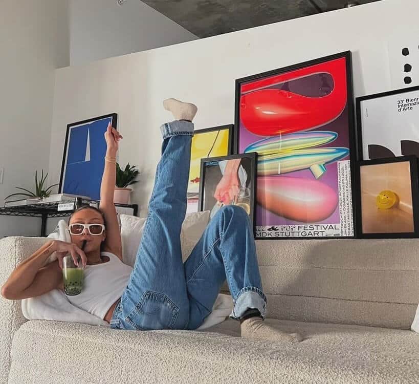 girl lying on the sofa drinking a smoothy with her legs in the air and a large gallery wall of artworks behind her