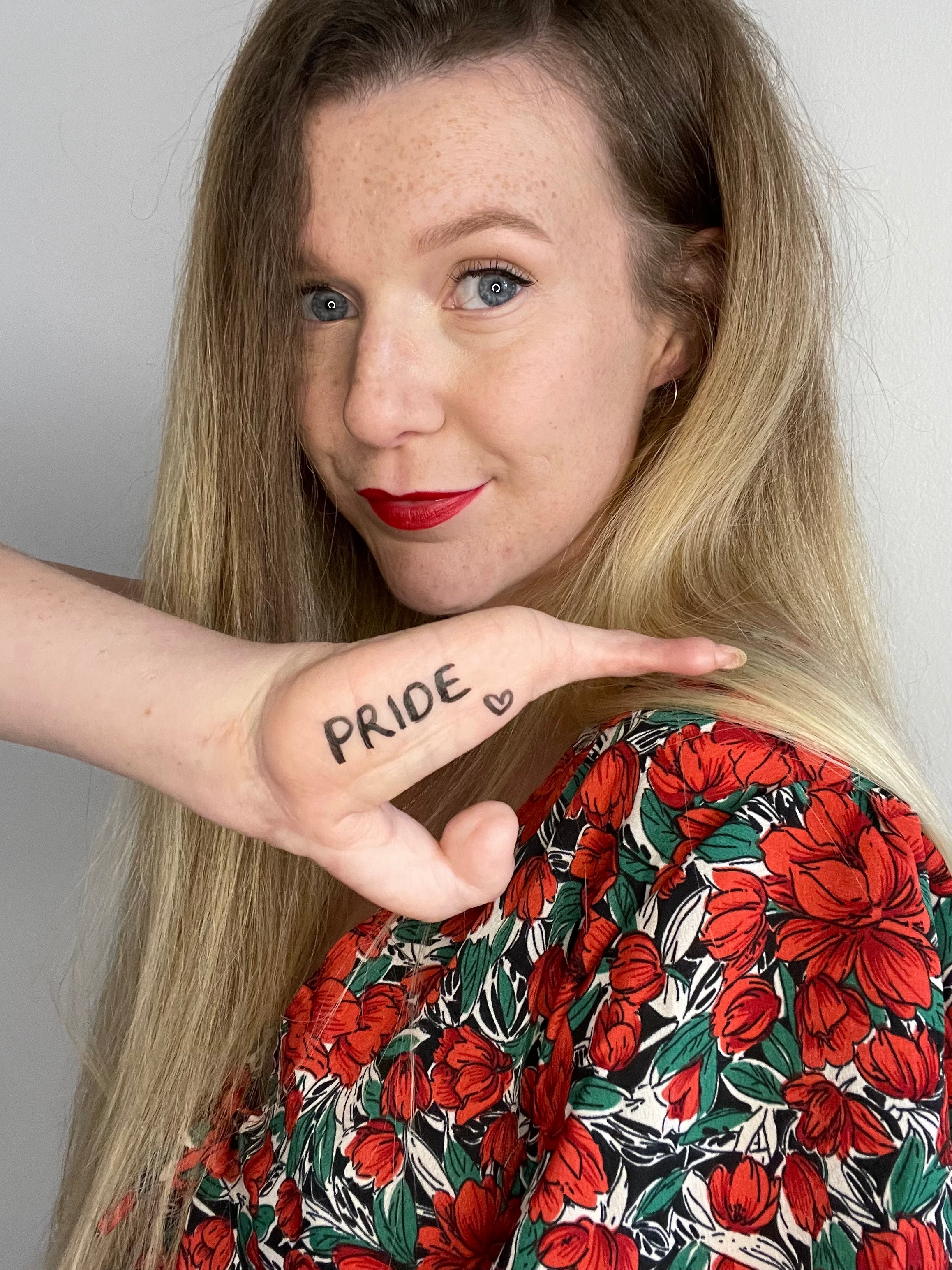 A woman with blonde hair and an upper limb difference is smiling at the camera with her hand up. Written on it says pride. 