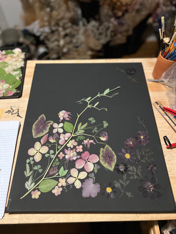 A black canvas is shown with pressed flowers on a major portion of it. The bottom 2/3 has lighter flowers and plant leaves and the bottom 1/3 has darker ones.