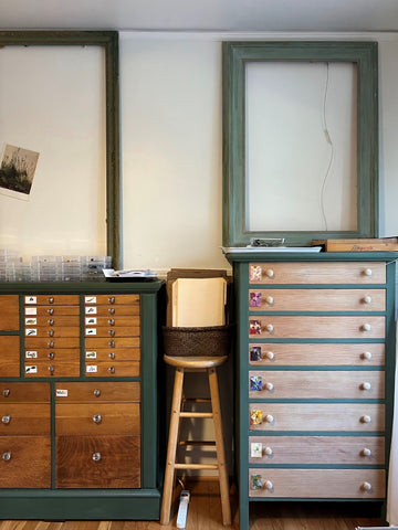 Two dressers with many drawers are side by side up against a wall. Empty frames which are as large as the dressers are resting on top of the cabinets. A stool sits between the dressers. Each drawer is labeled for a specific type of flower.