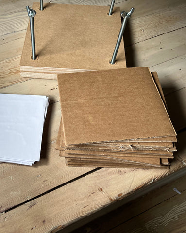 an image of a wood press with bolts, a stack of cardboard and a stack of papers, all siting on a wood floor.