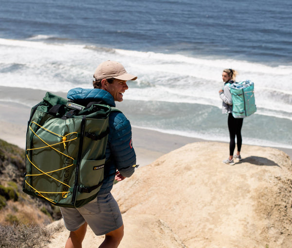 Man and woman hiking up a rocky coastline carrying their ULTRA standup paddleboards in backpacks