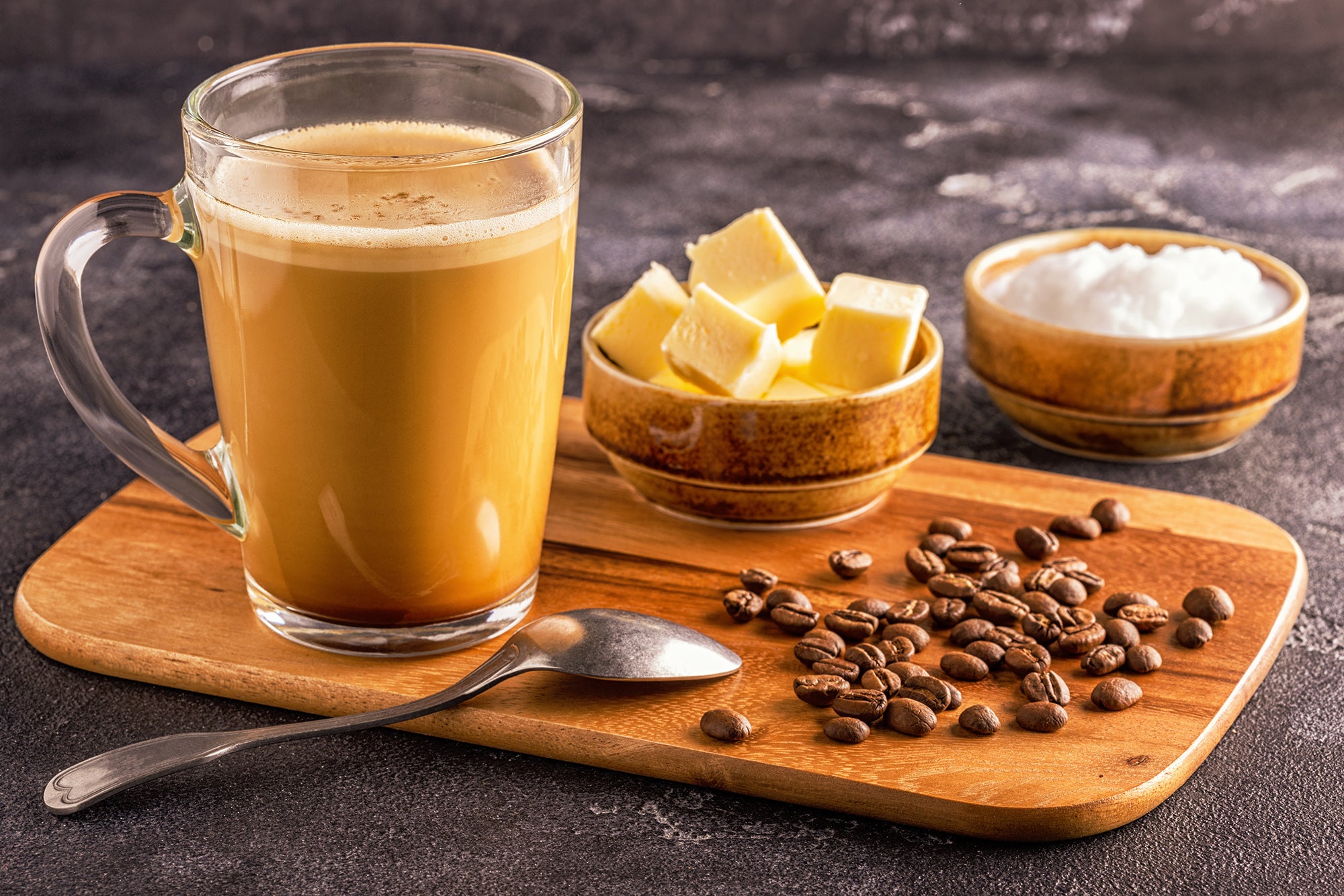 Coffee in a glass on a wooden board with a bowl of butter cubes and a bowl of coconut