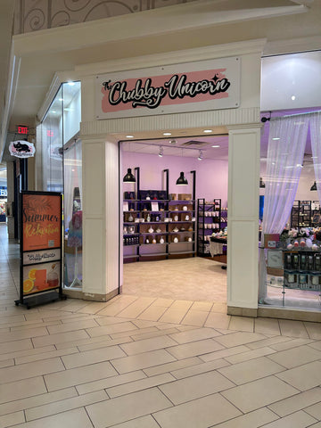 Direct front of the store in the Acadiana Mall. White outer walls with large glass windows to see inside. The chubby unicorn logo above the door. You can see the light pink walls inside.