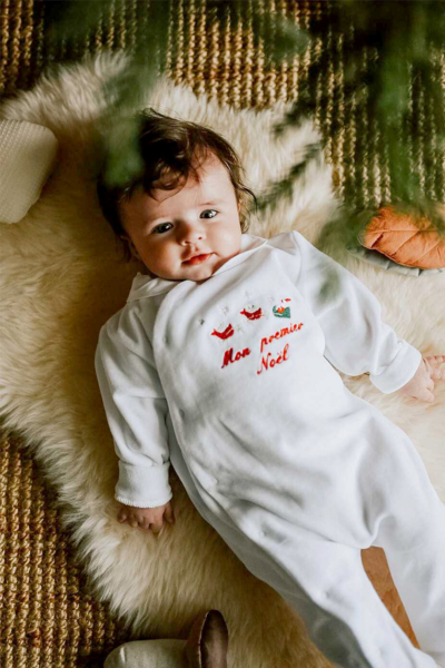 bébé qui porte un pyjama blanc avec brodé dessus en rouge 'mon premier noël'