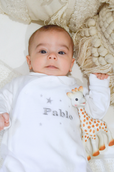 Bébé allongé sur un lit qui porte un pyjama blanc en velours de chez Bobine Paris avec 'Pablo' et des petites étoiles grises brodés dessus