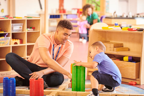 Male preschool teacher sitting on floor and helping a child build with colorful blocks.