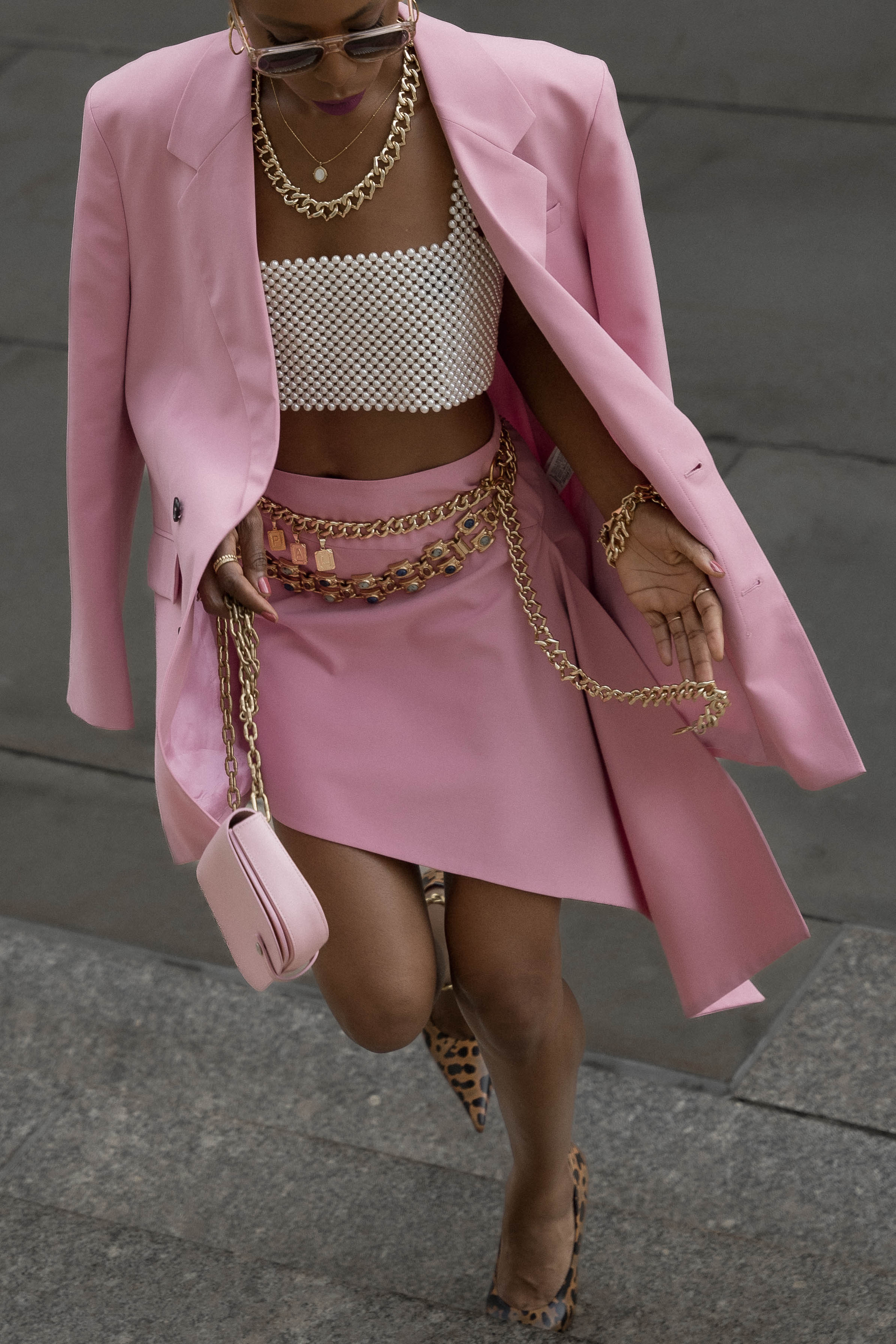 Photo of a woman on the street wearing pink skirt suit styled with pearl top, gold statement chain belt and jewelry