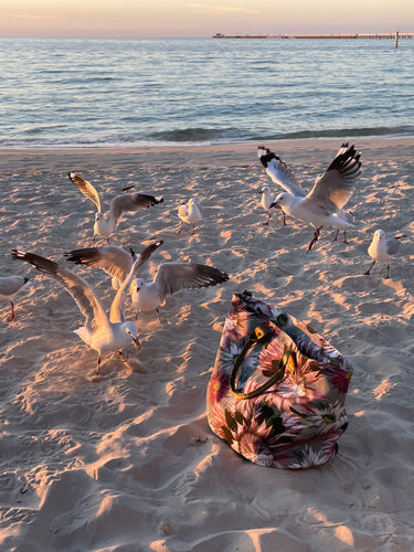 large water resistant, wildflower, canvas tote bag on the beach surrounded by flying seagulls. It is evening time and the sea is calm and the sand is full of footprints.