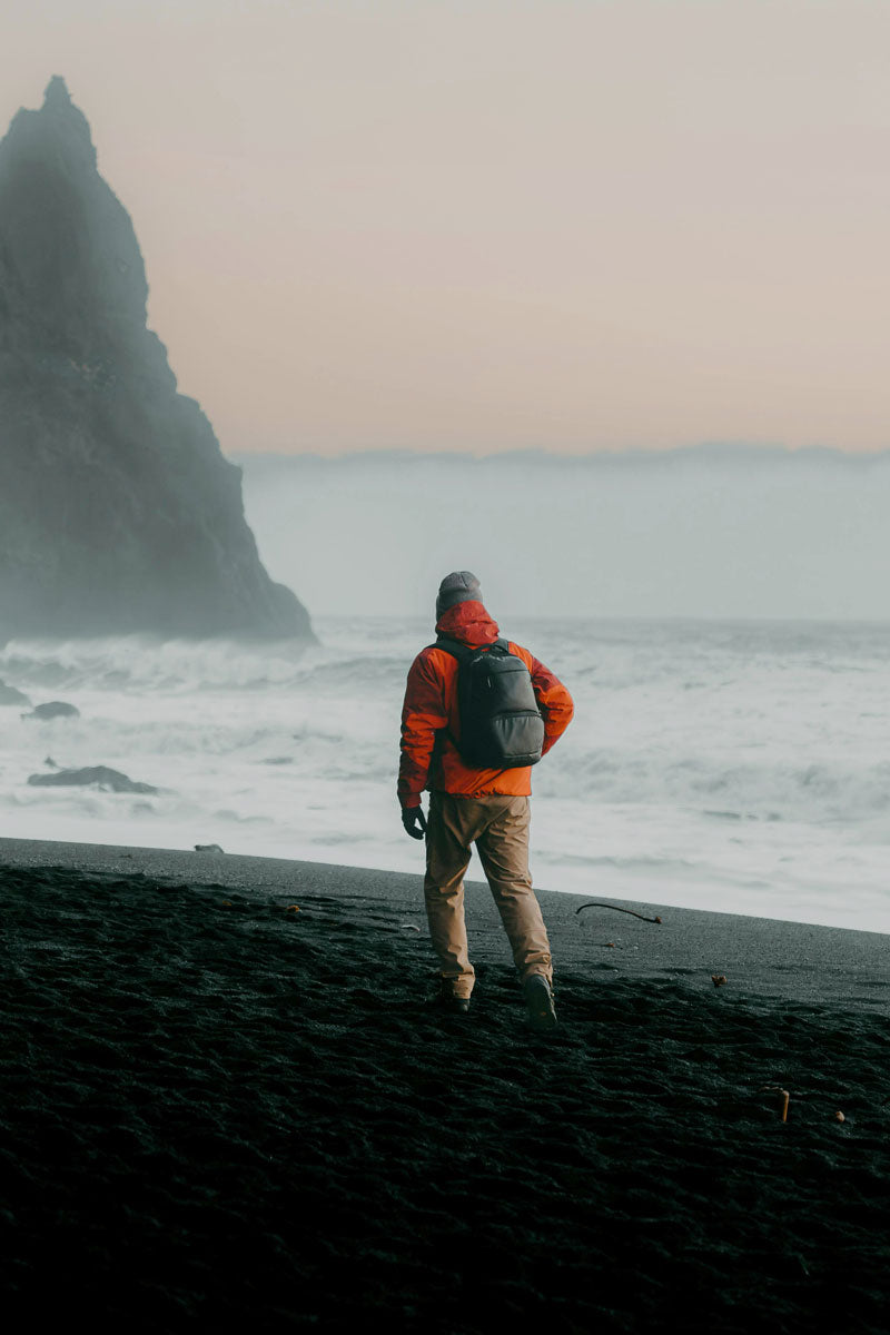 Person in an orange jacket and backpack walking on a foggy black sand beach.