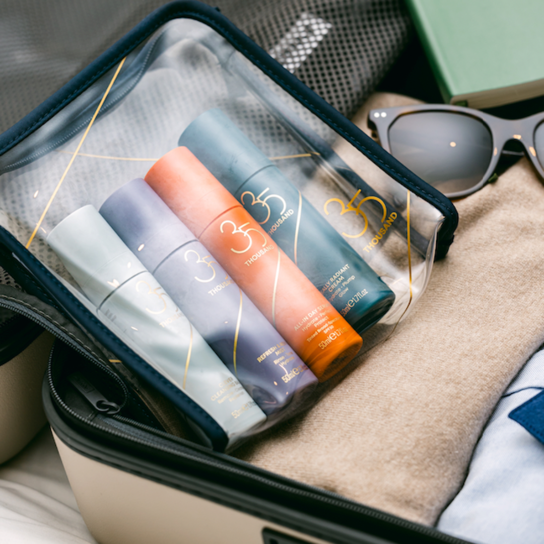 35 Thousand Signature Clear Travel Bag with Clever Cleansing Balm, Refresh & Revive Mist, All-In Day Serum with SPF 30 and Really Radiant Cream inside. Travel bag sitting atop an open piece of luggage with clothing and sunglasses, ready to be packed
