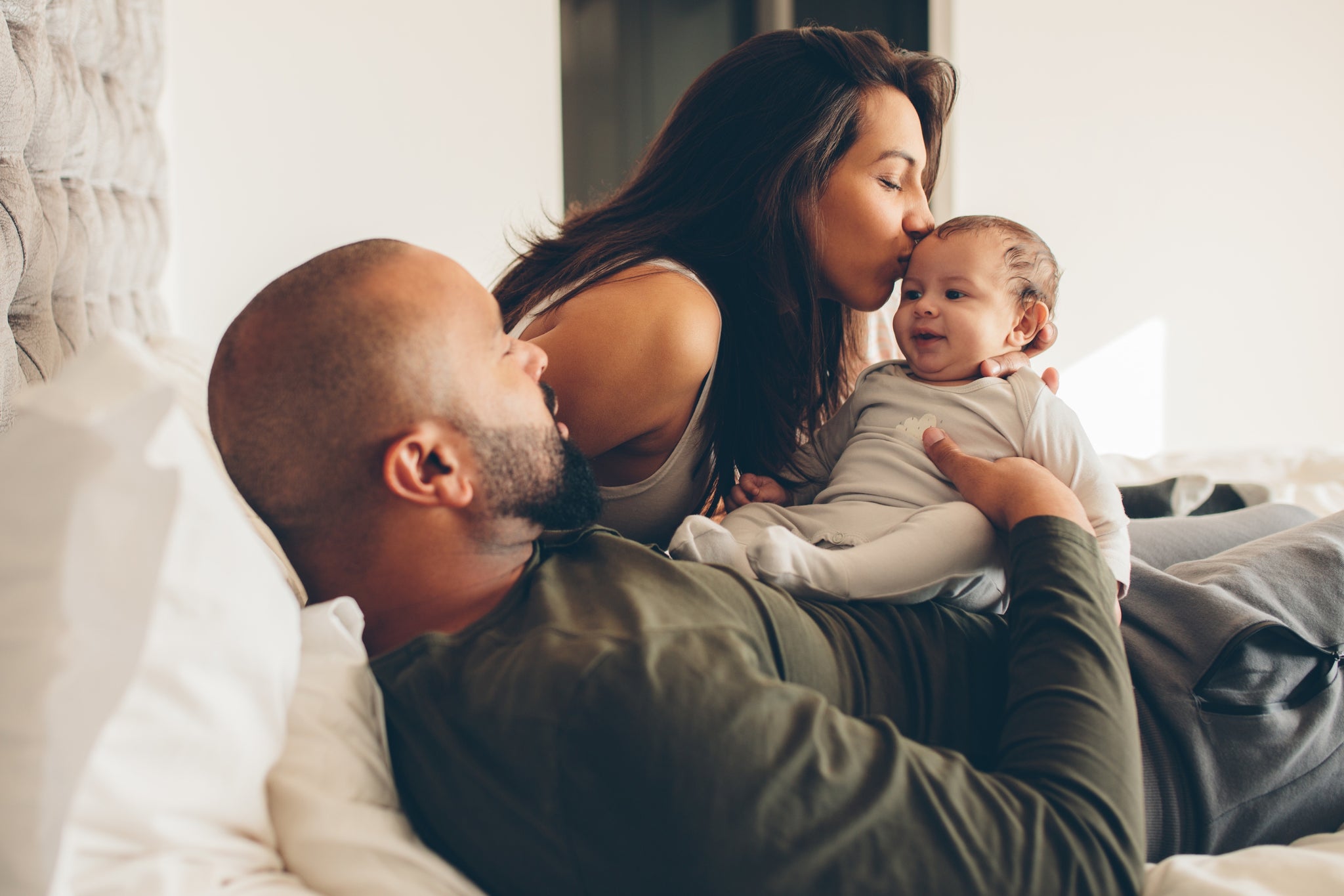 Father and mother on bed, holding baby. Father is lying on back and mother is kissing baby's head.