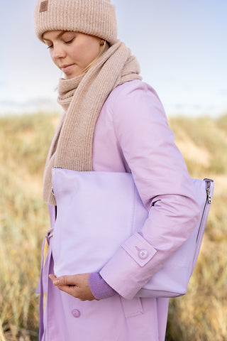 Nainen autiolla hiekkarannalla violetti takki päällä Utuan violetti käsilaukku kädessään.