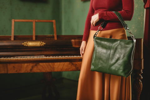 Kultamekkoinen nainen nojaa antiikkipianoon vihreä laukku kädellään.