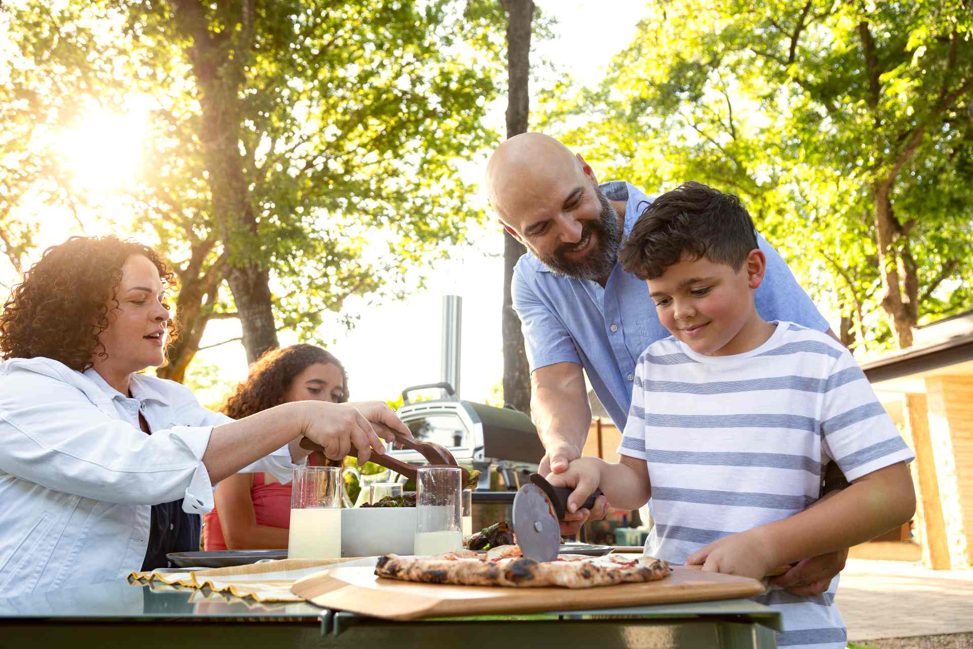 Man helping young boy cut a cooked pizza on an Ooni Bamboo Peel & Serving Board with an Ooni Professional Pizza Cutter Wheel with a woman serving salaf and a young girl next to an Ooni Karu 16.