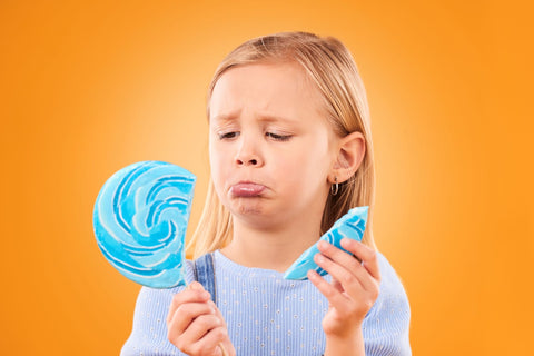A young girl looks sadly at her spoiled and broken blue-striped lollipop as she stands against an orange background.