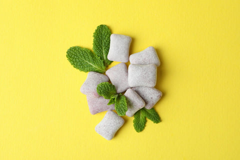 A close-up of chewing gums with mint leaves inserted between pieces, resembling a plant, set against a vibrant yellow background.