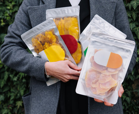 A girl, wearing a gray jacket over a black and white inner garment, holds packs of freeze-dried pineapple and mango; her face is not visible,