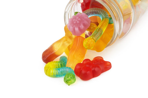 Neon gummy worms and assorted gummies in various shapes overflowing from a jar onto a surface.