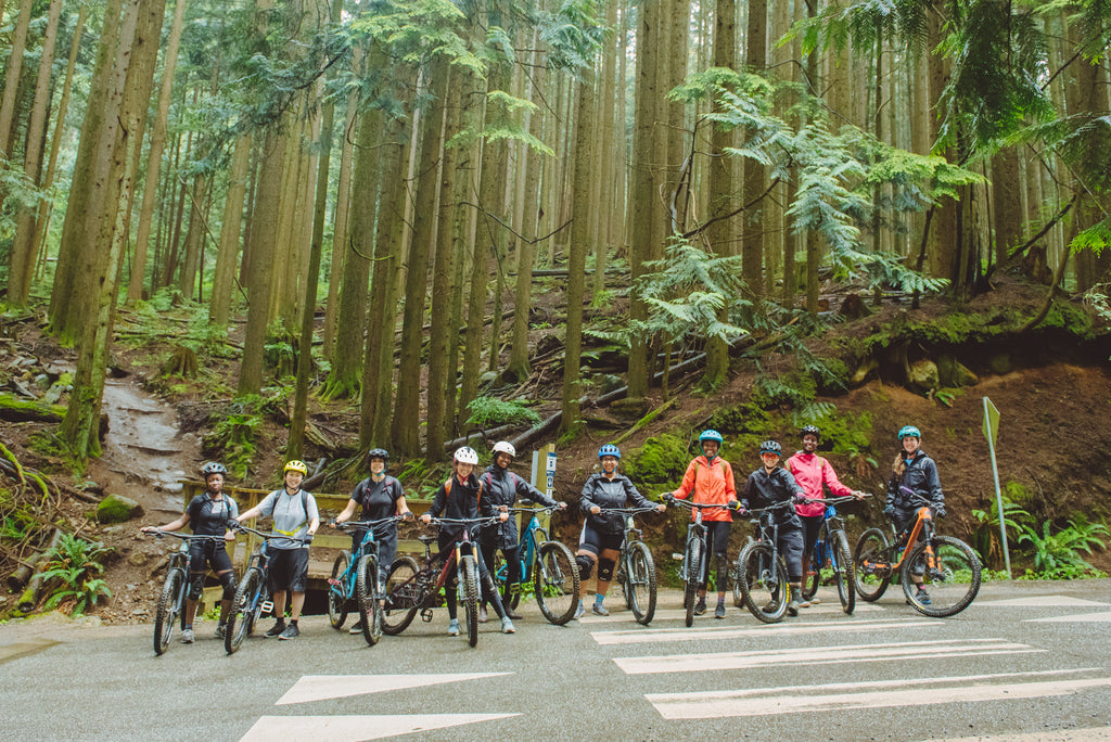 Group of BIPOC persons and allies stand with mountain bikes at the start of a forested trail ride