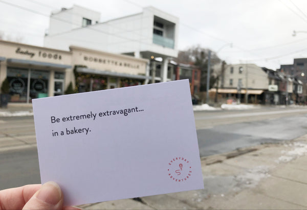 An Everyday Adventure prompt card reading "Be extremely extravagant...in a bakery" is held up in front of a bakery in the background